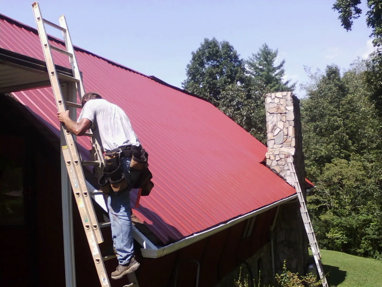 Roofer on a ladder beside a red metal roof and stone chimney — Soffit & Fascia Repair by Roofing Brothers in Avery Island